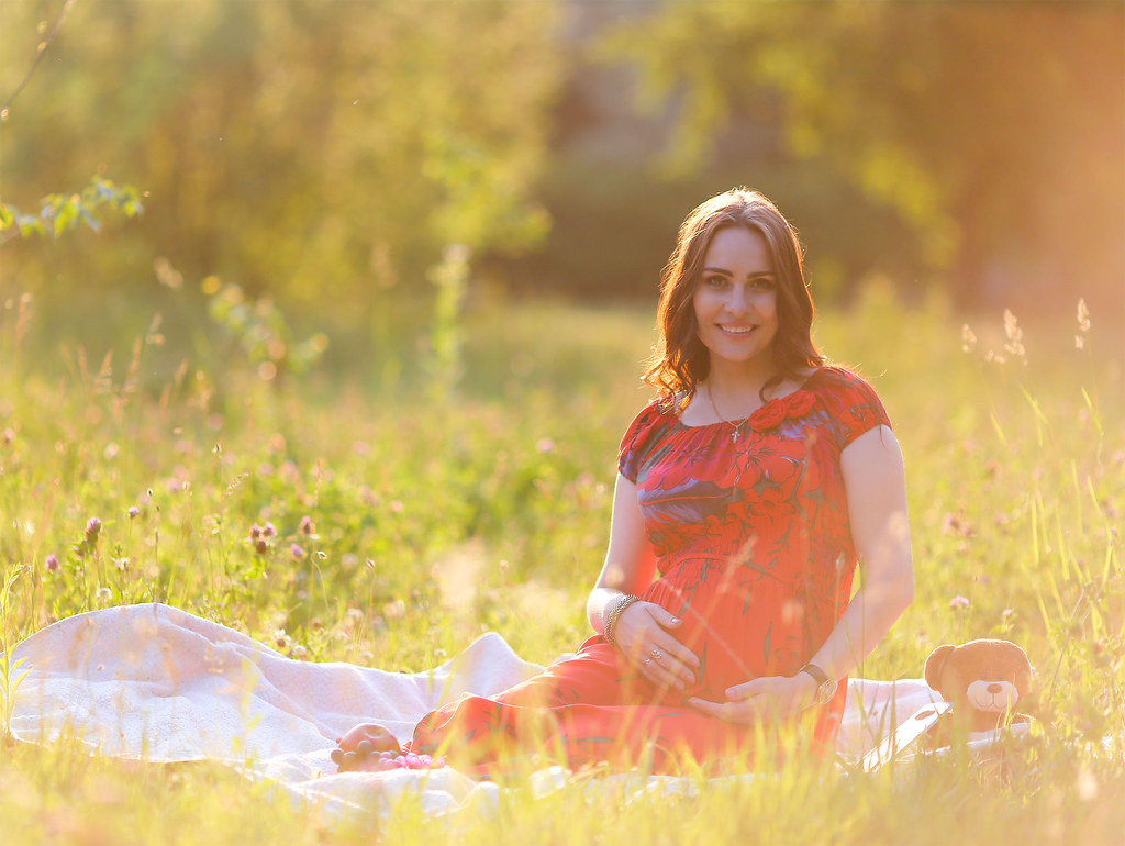 a pregnant woman in a red dress sits in grass on a sunny day. she is smiling and holding her stomach